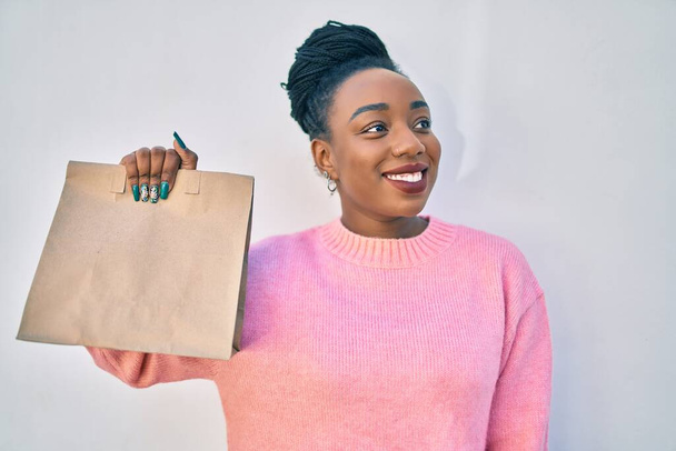 Junge Afroamerikanerin lächelt glücklich und hält Papiertüte mit Lebensmitteln zum Mitnehmen in der Stadt. - Foto, Bild