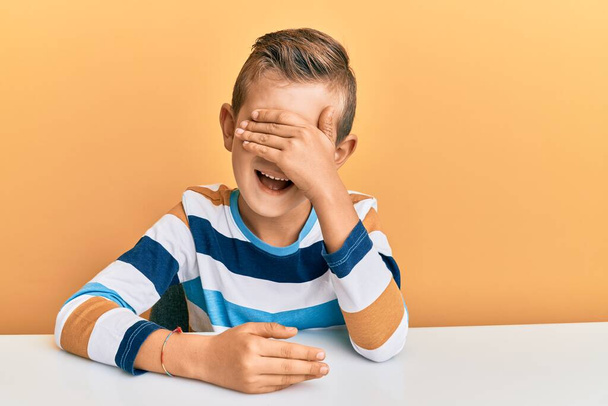 Adorable caucasian kid wearing casual clothes sitting on the table smiling and laughing with hand on face covering eyes for surprise. blind concept.  - Fotoğraf, Görsel