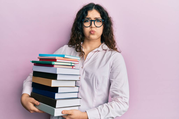 Young brunette woman with curly hair holding a pile of books puffing cheeks with funny face. mouth inflated with air, catching air.  - Photo, Image