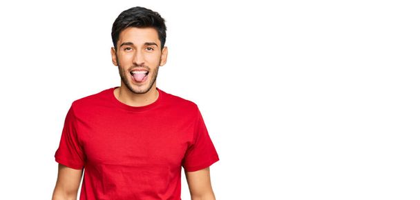 Kırmızı tişört giyen genç yakışıklı adam komik bir ifadeyle dilini dışarı çıkartıyor. duygu kavramı.  - Fotoğraf, Görsel