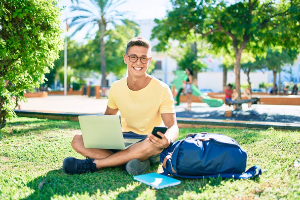 Mladý hispánský student usměvavý šťastný pomocí notebooku sedí na trávě na univerzitě kampusu - Fotografie, Obrázek