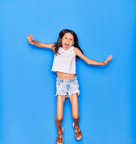 Entzückendes hispanisches Mädchen in lässiger Kleidung, das glücklich lächelt. Springen mit einem Lächeln im Gesicht über einen isolierten blauen Hintergrund - Foto, Bild