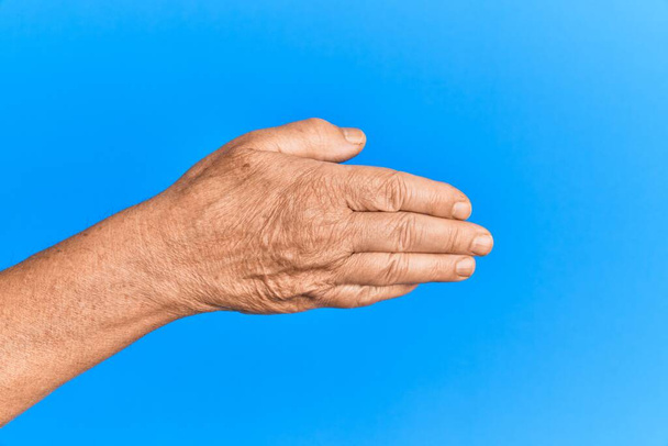 Ręka starszego Hiszpana nad niebieskim odizolowanym tłem rozciągającym się i sięgającym z otwartą ręką do uścisku dłoni, pokazującym tył dłoni  - Zdjęcie, obraz