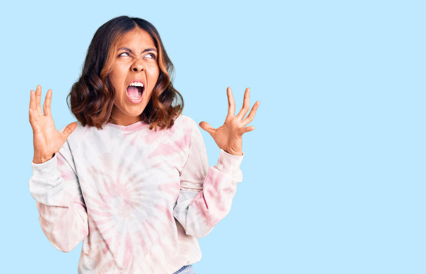 Young beautiful mixed race woman wearing casual tie dye sweatshirt crazy and mad shouting and yelling with aggressive expression and arms raised. frustration concept.  - Фото, изображение