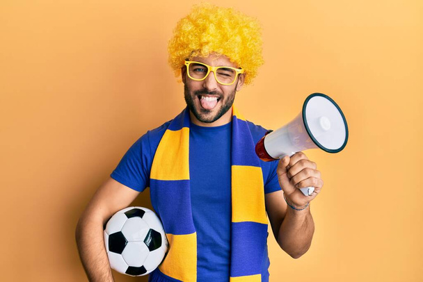 Giovane uomo ispanico tifoso di calcio che tiene palla da calcio utilizzando megafono attaccare lingua fuori felice con espressione divertente.  - Foto, immagini