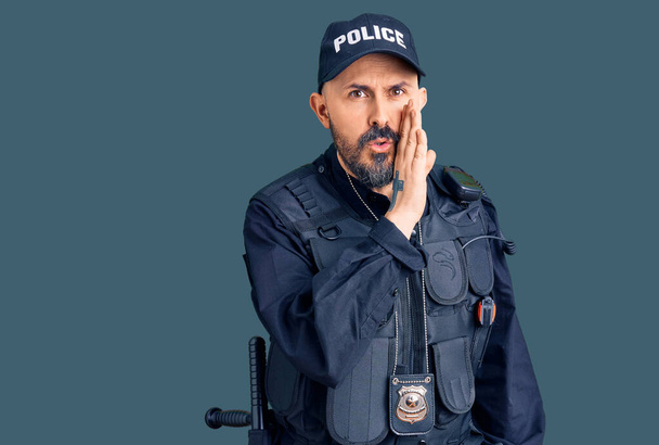 Joven hombre guapo vistiendo uniforme de policía mano en boca contando rumores secretos, susurrando conversación maliciosa  - Foto, imagen
