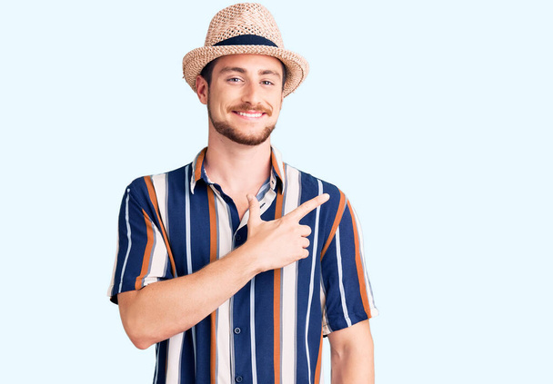 Young handsome caucasian man wearing summer hat cheerful with a smile of face pointing with hand and finger up to the side with happy and natural expression on face  - Foto, imagen