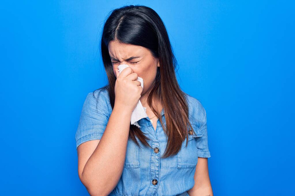 Jonge mooie vrouw ziek van de griep. Met behulp van papieren zakdoek op de volle neus van slijm staande over geïsoleerde blauwe achtergrond - Foto, afbeelding