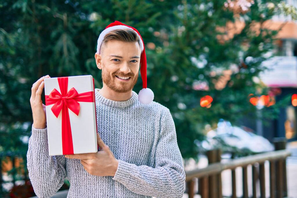 Jeune homme caucasien portant un chapeau de Noël tenant un cadeau au parc. - Photo, image