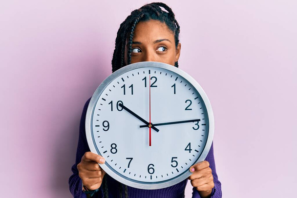 Африканська американка з плечима, що тримає великий годинник закриваючи обличчя посміхаючись дивлячись з боку в бік і дивлячись геть думки.  - Фото, зображення