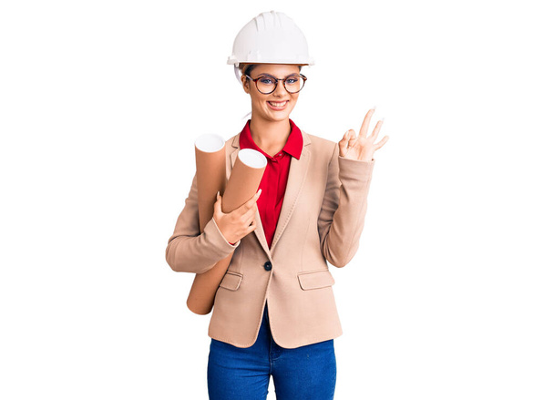 Młoda piękna kobieta nosi architekt hardhat i okulary trzymając plany robi ok znak palcami, uśmiechając przyjazny gest doskonały symbol  - Zdjęcie, obraz