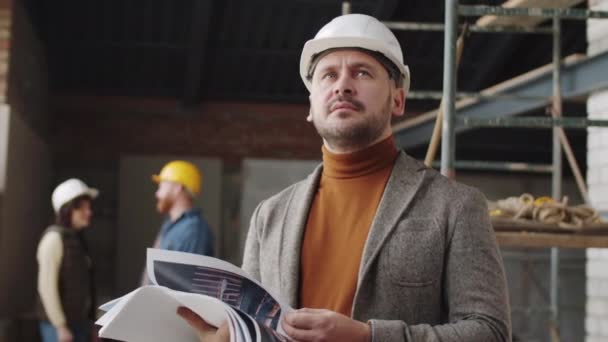 Pas nahoru záběr zamyšleného mužského inženýra v klobouku zkoumá stavební plán na papíře a rozhlíží se po staveništi - Záběry, video