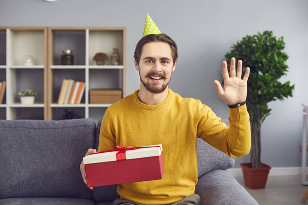 Smiling man with holiday present box in hand looking at camera and waving during videocall - Foto, imagen