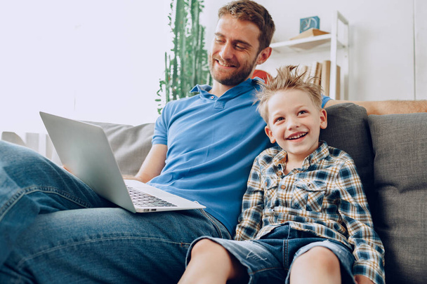 Father and son sitting on couch and watching something on laptop - Фото, изображение