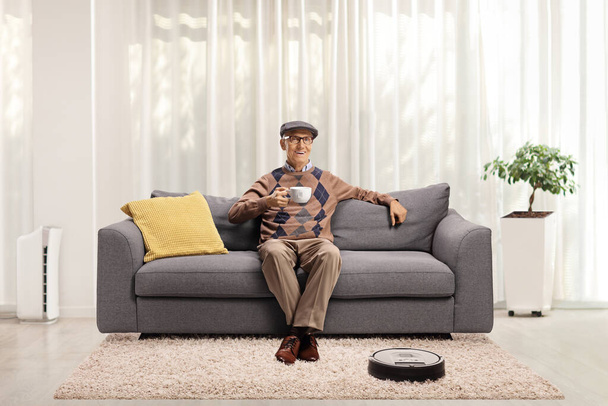 Robotic vacuum cleaner cleaning the floor and an elderly man drinking tea on a sofa at home - Photo, Image