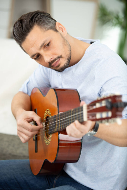 pretty young man playing guitar while sitting on sofa - Фото, зображення