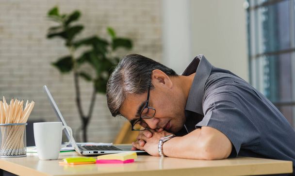Asian businessman tired overworked he sleeping over a laptop computer on the desk. senior man with eyeglasses lying asleep on table at his working place - 写真・画像