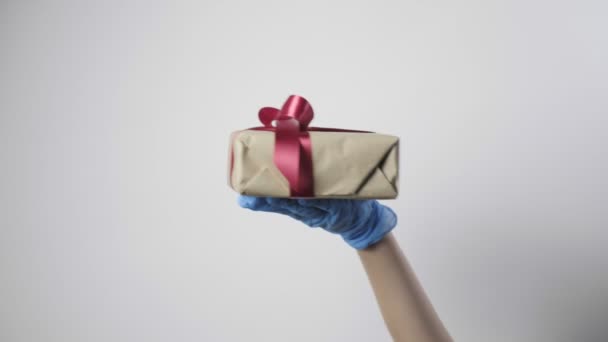 Hand in blauem Schutzhandschuh, Geschenkbox mit Schleife. COVID-19 Neujahr, Weihnachten, Valentinstag-Überraschung. Quarantäne-Sicherheit, neue Normalität. Kind schenkt Geschenk auf weißem Hintergrund - Filmmaterial, Video