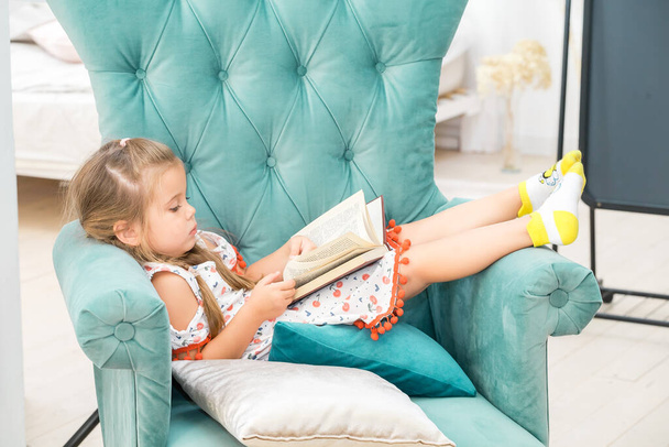 A little girl lies in a green chair and reads a large heavy book. - 写真・画像