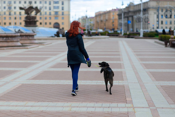 Young female using a face mask as a coronavirus spreading prevention walking with her dog. Global COVID-19 pandemic concept image. - Photo, image