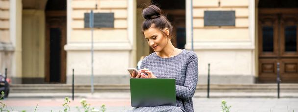 Nuori nainen istuu nurmikolla matkapuhelimen ja kannettavan tietokoneen kanssa yliopiston taustaa vasten - Valokuva, kuva