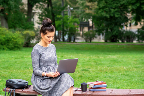 Όμορφη νεαρή γυναίκα κάθεται σε ένα παγκάκι με ένα φορητό υπολογιστή και εργάζεται ή σπουδάζει σε εξωτερικούς χώρους - Φωτογραφία, εικόνα