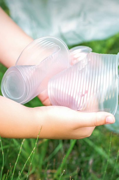 Τα χέρια του παιδιού καθαρίζουν το πάρκο από χρησιμοποιημένα πλαστικά σκεύη στο γρασίδι - Φωτογραφία, εικόνα