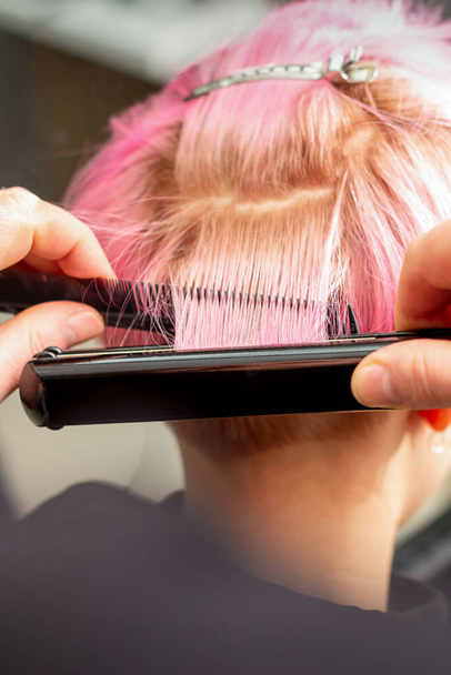 Vista da vicino sulle mani del parrucchiere raddrizzando i capelli rosa corti con una piastra per capelli in un salone di parrucchiere - Foto, immagini