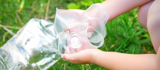 Τα χέρια του παιδιού καθαρίζουν το πάρκο από χρησιμοποιημένα πλαστικά σκεύη στο γρασίδι - Φωτογραφία, εικόνα
