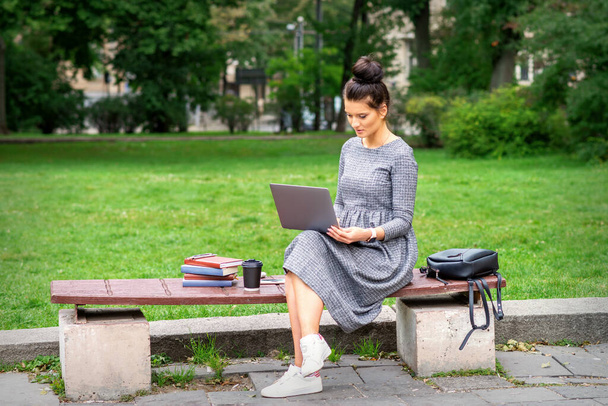 Beautiful young woman sitting on a bench with a laptop and working or studying outdoors - Foto, Bild