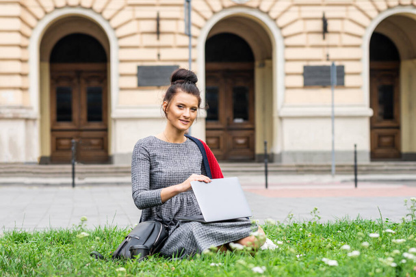Egy gyönyörű, fiatal, mosolygó diák portréja, aki laptoppal a füvön tanul az egyetemi épület mellett. - Fotó, kép