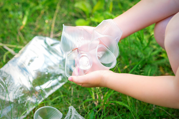 Child's hands clean the park from used plastic utensils in the grass - Photo, Image