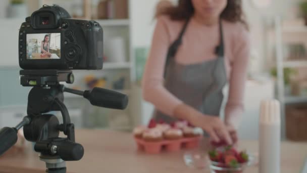 Kamera auf Stativ filmt, wie sie in der Küche vor einer verschwommenen Frau steht, die Cupcakes mit Beeren dekoriert - Filmmaterial, Video