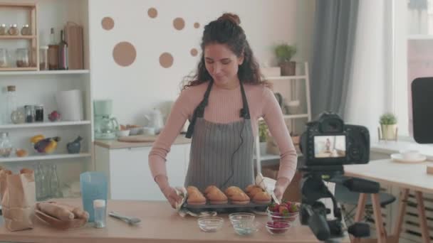 Tracciamento medio sinistro della giovane donna caucasica in grembiule in piedi a tavola, mettendo una teglia da forno con muffin sopra, parlando e sorridendo sulla macchina fotografica montata su treppiede di fronte a lei - Filmati, video
