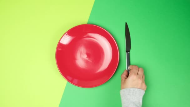 női kéz fel egy zöld háttér fém villa és kés, mellett egy üres piros kerek kerámia lemez, felülnézet - Felvétel, videó