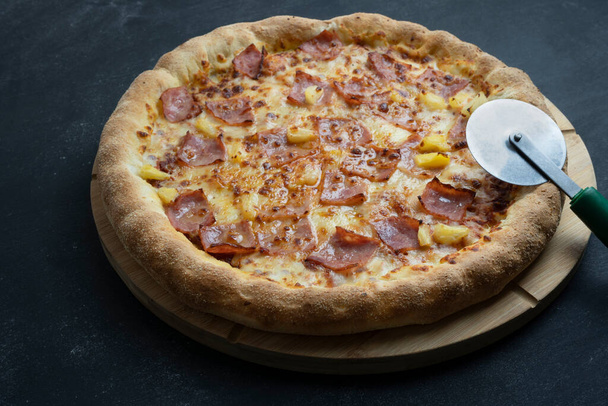 Maukas havaijilainen pizza (juusto, rapea pekoni ja ananas) ja pizzaleikkuripyörä.Tumma tausta Kopioi tilaa . - Valokuva, kuva