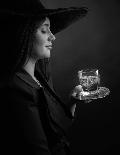 Attractive girl with alcoholic drink in frozen glass. Beautiful brunette in black hat holding glass with cold cocktail. Black and white. - Photo, Image