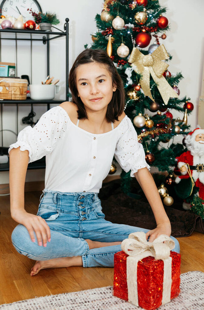 Beautiful teenage girl 12-year old sitting with Christmas presents under Christmas tree. Sitting on floor in room - Zdjęcie, obraz