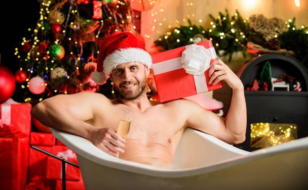 Sorry Santa, naughty just feels nice. Christmas gift. Sexy bearded man relaxing in hot bath with champagne and gift box. Xmas gift. Man muscular torso lying in bathtub winter decorations background - Фото, изображение