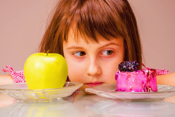 Молодая милая красивая девушка, имеющая жесткий выбор между здоровой и нездоровой пищи. Она выбирает между яблочными фруктами и вкусным тортом.  - Фото, изображение