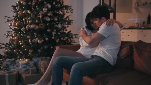 νεαρό ζευγάρι αγκαλιάζει και φιλιά στον καναπέ κοντά στο χριστουγεννιάτικο δέντρο - Πλάνα, βίντεο