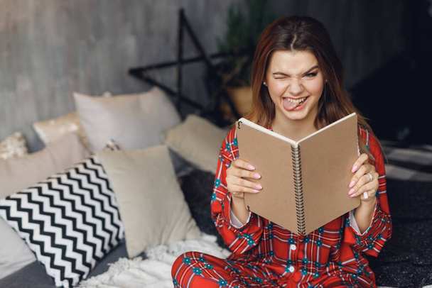 Close up of girl wears festive pajamas and take brown note in hands at home. Crazy woman with tongue in red night-wear on bed - Fotografie, Obrázek