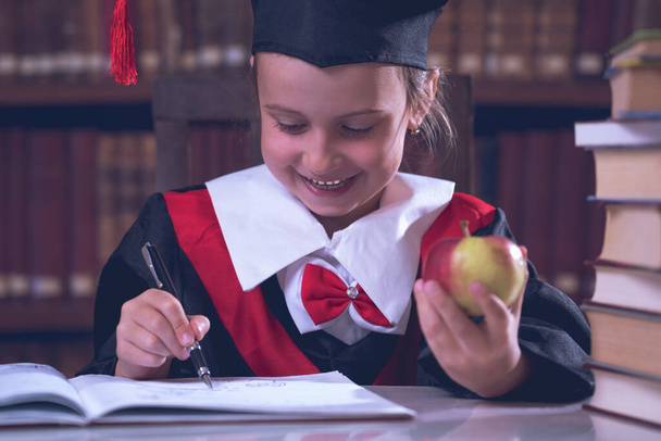 Eğitim konsepti. Mantodaki kız öğrenci kütüphanede kitap okuyor. Genç ve güzel bir kız öğrenim görüyor ve bilginin sembolü olarak elinde bir elma tutuyor. Yatay resim. - Fotoğraf, Görsel