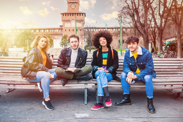 Grupo de cuatro multiétnicos mejores amigos sentado al aire libre banco al aire libre en Milán divertirse pasar tiempo juntos en buena compañía - Foto, imagen