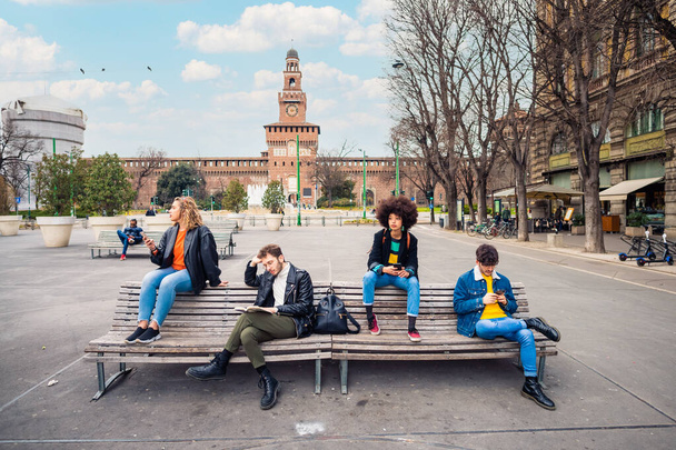 Młody mężczyzna kaukaski czyta książkę odróżniającą od innych wielonarodowych ludzi nieznajomi lub przyjaciele siedzący na ławce outdoor za pomocą smartfona ignorując uzależnionych od trendów społecznych  - Zdjęcie, obraz