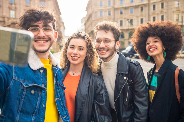 Grupo de amigos multiétnicos al aire libre divertirse y tomar selfie utilizando el teléfono inteligente al aire libre en la ciudad sonriendo feliz con vibraciones positivas al atardecer - Foto, Imagen