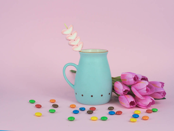 A blue ceramic mug with hot chocolate and marshmallows on a stick, with flowers and candy on a pink background - Фото, изображение