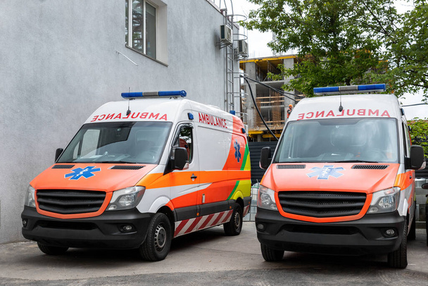 Dwa nowoczesne białe i pomarańczowe ambulans ambulans medyczny furgonetka zaparkowany w pobliżu budynku szpitala. Sanitariusze na ulicy. Opieka zdrowotna i koncepcja pilnego wsparcia medycznego - Zdjęcie, obraz