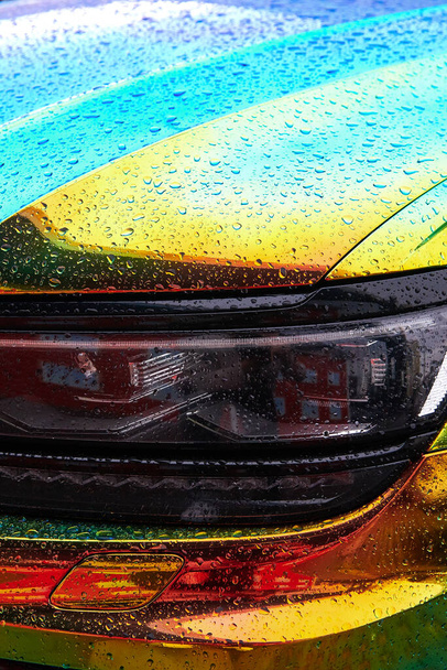 Coche de color holográfico camaleón bajo la lluvia. Envoltura del coche con pintura iridiscente. Gotas de lluvia en el coche. Vista frontal de cerca. Faros led modernos. Envoltura del coche. Foto vertical. - Foto, Imagen