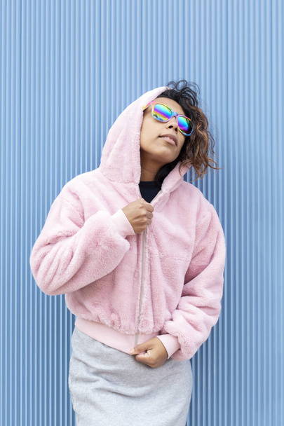 Portrait of a young hispanic woman wearing sunglasses with the colors of the lgbt flag. She is standing isolated on a blue background. Concept of sexual freedom and diversity. - Fotografie, Obrázek